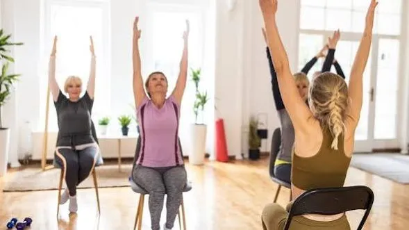Older adults seated on chairs in a bright room, arms raised overhead